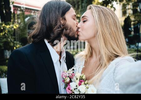 Junge schöne Bräutigam und Braut sinnlich küssen während der Aufnahme Selfie im Garten des Restaurants Stockfoto