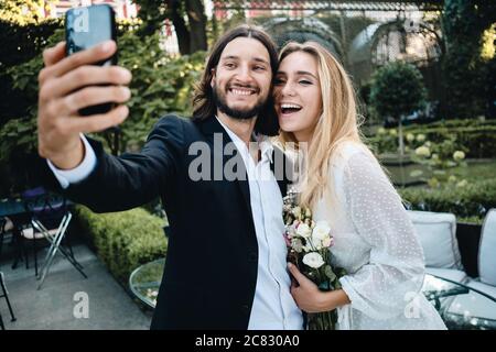 Junge attraktive Bräutigam und Braut freudig unter Selfie auf Handy in schönen Garten des Restaurants Stockfoto