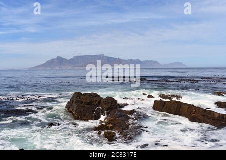 Tafelberg, Lions Head & Devils Peak von Robben Island aus gesehen, gegenüber der Table Bay. Der Ozean stürzt gegen die felsige Uferlinie. Stockfoto
