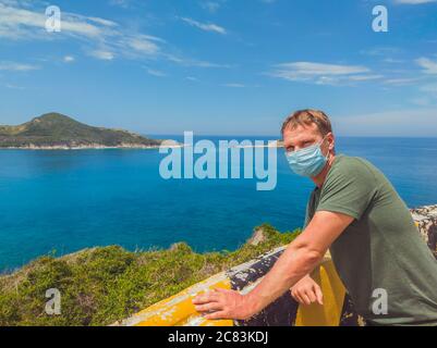 Junge schöne Mann schützen vor Luftverschmutzung oder Coronavirus durch das Tragen einer medizinischen Maske, zu Fuß außerhalb im Park in der Nähe azurblauen Meer, blauen Himmel. Stopp Stockfoto