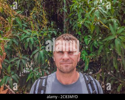 Ein schöner junger Kaukasier in einem grauen Hemd mit Rucksack in der Nähe von grünem Mango Baum. Reisekonzept mit Kopierplatz Stockfoto