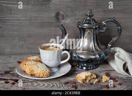 Schwarzer Kaffee in einer stilisierten viktorianischen Tasse und Haferflocken-Kekse, die mit Sesam auf einem Porzellanteller bestreut wurden, sowie alte silberne Kaffeekanne und Gra Stockfoto