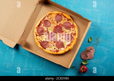 Traditionelle italienische Pepperoni oder Salami-Pizza im Ofen in einer Pappschachtel, die zum Mitnehmen bereit ist, mit frischen Zutaten auf blauem Holz in einer Ove Stockfoto