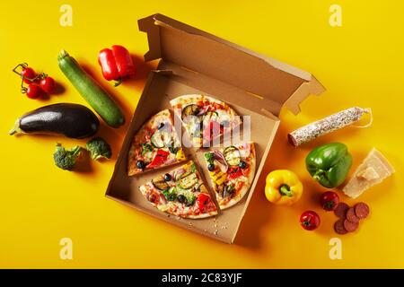 Köstliche italienische Pizza mit frischem Gemüse und Salami Topping in Portionen in einer Take-away-Box geschnitten, flankiert von verschiedenen frischen Zutaten auf einem gelben Stockfoto