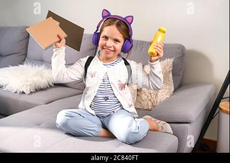 Kleine Mädchen musikalische Kopfhörer machte sich bereit, zur Schule zu gehen. Schulmädchen mit Rucksack, Büchern und einem Snack für den Schultag. Bildung, Lernkonzept. Glückliches Kind. Freier Speicherplatz für Text Stockfoto