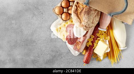 Verschiedene Milchprodukte mit Eiern, Pasta und Salami, die aus einem wiederverwendbaren Einkaufstasche auf einen strukturierten grauen Hintergrund mit Copyspace in einem Panorama verschüttet werden Stockfoto