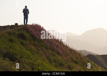 Ein junger Tourist mit Rucksack bewundert den Sonnenuntergang in den Bergen vor dem Hintergrund blühender Hänge. Entfernte Bergketten wi Stockfoto