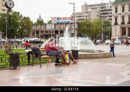 Iasi / Rumänien - 14. Mai 2020: Erwachsene Männer sitzen auf der Parkbank neben dem Brunnen und trinken Alkohol, während der Junge mit dem Wasserpistole spielt Stockfoto