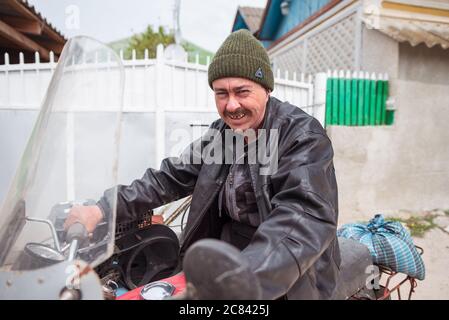Trebujeni / Moldawien - 15. Mai 2020: Moldawischer Mann mit Goldzähnen und Wollmütze lächelte die Kamera an, während er in einem Dorf auf einem alten Motorrad fuhr Stockfoto