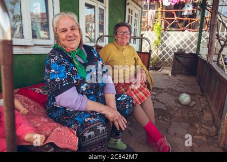 Trebujeni / Moldawien - 15. Mai 2020: Portrait von zwei erwachsenen Frauen, die auf der Veranda ihres Hauses sitzen und traditionelle Kleidung tragen, im ländlichen Raum Moldawiens, die vor der Kamera lächeln Stockfoto
