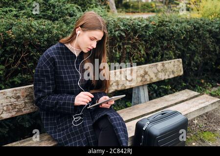 Schöne Casual Mädchen in Ohrhörer nachdenklich mit Tablet sitzen mit Koffer auf Bank im Park Stockfoto