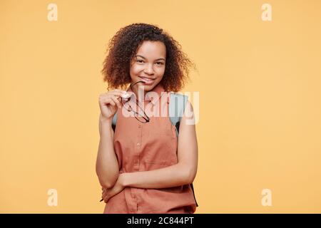 Porträt einer lächelnden jungen afroamerikanischen Frau, die eine Brille neben dem Mund hält und vor gelbem Hintergrund posiert, Studentenkonzept Stockfoto