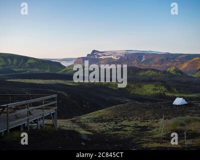 Holzterrasse und weißes Zelt auf Botnar Campingplatz bei Island auf Laugavegur Wanderweg, grünes Tal in vulkanischer Landschaft mit Blick auf Myrdalsjokull Stockfoto