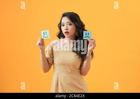 Emotionale Frau hält Notizbuch mit gezeichneten Emoticons auf gelbem Hintergrund Stockfoto