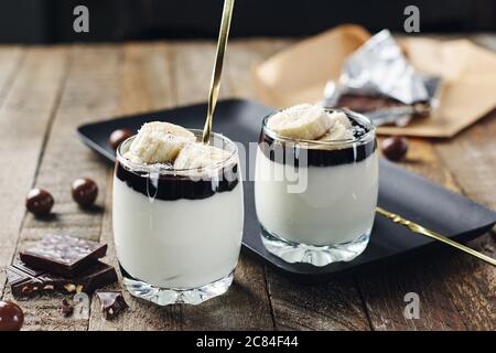 Milchdessert mit Banane und Schokoladencreme in einem Glas auf Holzgrund. Stockfoto