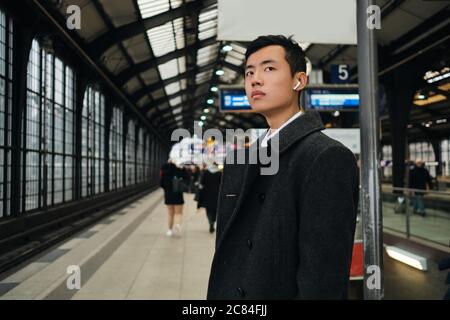 Junger, stilvoller asiatischer Geschäftsmann in schnurlosen Kopfhörern, der am Bahnhof in der Bahn wartet Stockfoto
