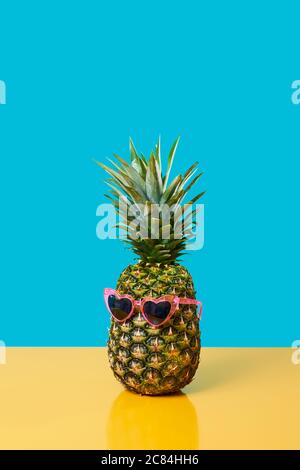 Eine Ananas mit einer rosa herzförmigen Sonnenbrille auf einer gelben Oberfläche auf einem blauen Hintergrund mit etwas leerem Platz oben Stockfoto