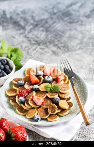 Zum Frühstück gab es winzige Pfannkuchen. Cereal Pancakes mit Heidelbeeren, Erdbeeren auf Teller auf grauem Hintergrund. Trendiges Essen. Platz für Text oder Design kopieren Stockfoto