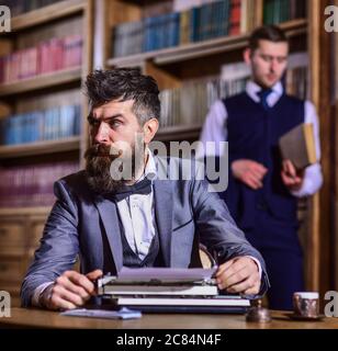 Portrait von stilvollen schönen Schriftsteller mit Bart. Mann in Anzug und Fliege, sitzt neben Retro-Schreibmaschine vor der Bibliothek oder Vintage-Interieur, defokussed Stockfoto