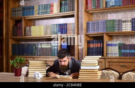 Wissenschaft und Studium Konzept. Forscher oder Lehrer Buch lesen. Mann im klassischen Anzug oder Professor mit enthusiastischem Gesicht sitzt in der Bibliothek in der Nähe von Bücherregalen auf dem Hintergrund, entkokelt. Stockfoto