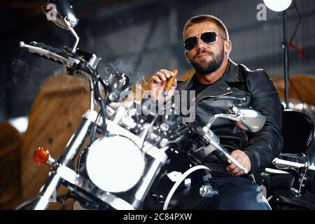 Cool bärtiger Mann Biker in Sonnenbrille auf seinem Motorrad sitzen und Zigarre rauchen Stockfoto