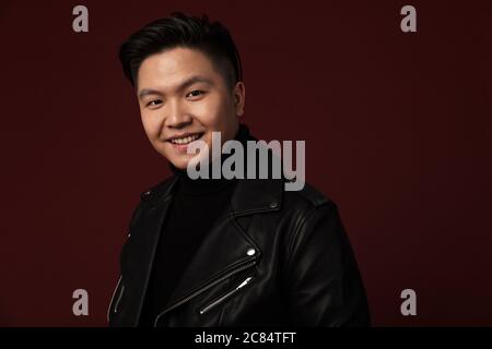 Bild von fröhlichen asiatischen Mann in Lederjacke lächeln und Blick auf Kamera isoliert über Burgund Hintergrund Stockfoto