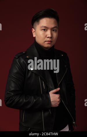 Bild von ernsthaften asiatischen Mann in Lederjacke posiert und Blick auf Kamera isoliert über Burgund Hintergrund Stockfoto