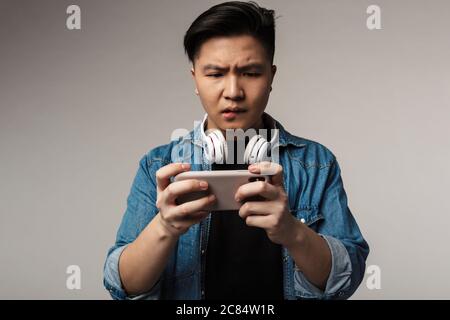 Bild von unglücklichen jungen asiatischen Mann spielen Videospiel auf Smartphone isoliert über grauen Hintergrund Stockfoto