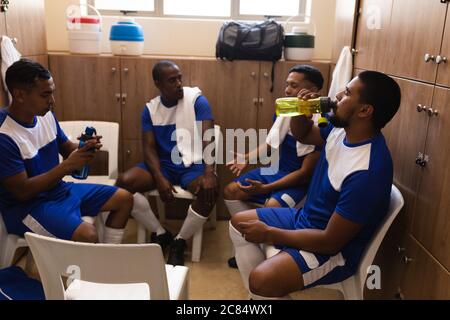 Multiethnische Gruppe von männlichen Fußballspielern, die einen Mannschaftsstreifen tragen, sitzen in der Umkleidekabinen während einer Spielpause, ruhen sich aus, halten und trinken Stockfoto