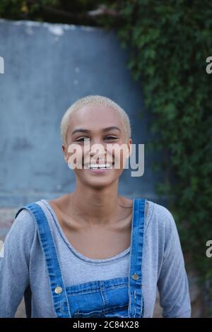 Portrait einer alternativen Mischrennerin mit kurzen blonden Haaren und Denim-Latzhose an einem sonnigen Tag in der Stadt, lächelnd vor der Kamera. Stadt Stockfoto
