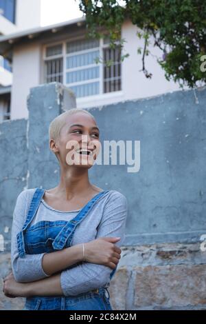 Gemischte Rasse alternative Frau mit kurzen blonden Haaren in Denim Latzhose, unterwegs in der Stadt an einem sonnigen Tag, wegschauen und lachen. Urba Stockfoto