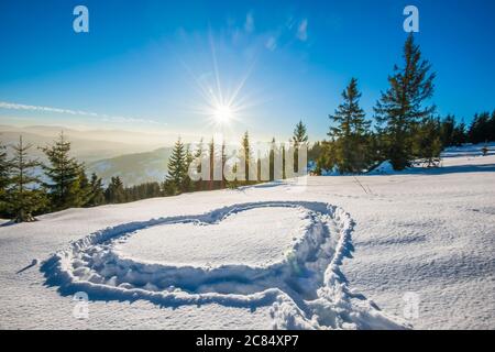 Herz trampelte auf dem Schnee mit Füßen in einer Schneeverwehung auf einem Hang mit herrlichem Blick auf den Nadelwald und die Bergketten auf einem sonnigen Frost Stockfoto