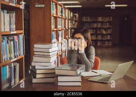 Gelangweilte junge Studentin, die am Tisch in einer offenen Bibliothek auf dem Campus sitzt. Bildungskonzept Stockfoto