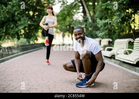 Schöner lächelnder afrikanischer Mann, der seine Schuhe schnürte und sich mit seiner Freundin, einer hübschen kaukasischen Frau, auf Laufen und Sport im Stadtpark vorbereitete Stockfoto