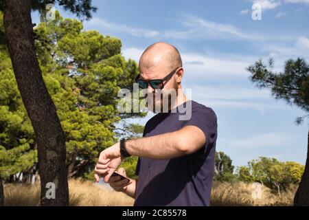 Junger Erwachsener, kahl und bärtig, trägt schwarze Sonnenbrille und steht an heißen Tagen in der Natur und überprüft seine Uhr Stockfoto