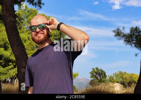 Junger bärtiger und kahler Erwachsener trägt eine Sonnenbrille und lächelt und genießt sich an einem warmen Tag draußen in der Natur Stockfoto