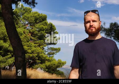 Junger bärtiger kahler Erwachsener mit Sonnenbrille auf dem Kopf, der draußen in der Natur steht und einen besorgten Ausdruck in die Ferne blickt Stockfoto