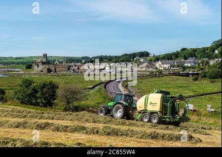 Timoleague, West Cork, Irland. Juli 2020. An einem Tag mit heißem, sonnigem Wetter und hohen 22 Grad Celsius, Michael und Richard McCarthy Ballensilage auf ihrer Farm in Timoleague, West Cork. Timoleague und Timoleague Abbey sonnen sich im Hintergrund. Quelle: AG News/Alamy Live News Stockfoto