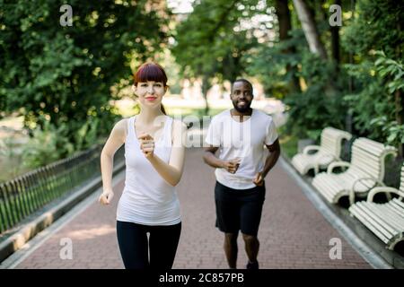 Athletisches multiethnisches Paar, dunkelhäutiger Mann und kaukasische rothaarige Frau, die zusammen laufen. Sportläufer joggen früh auf Parklandschaft Stockfoto