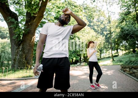 Sport Lifestyle Konzept. Junge starke afrikanische Mann müde nach harten Cardio-Training, stehen mit Wasserflasche. Erschöpft kaukasischen Mädchen ruhen nach Stockfoto