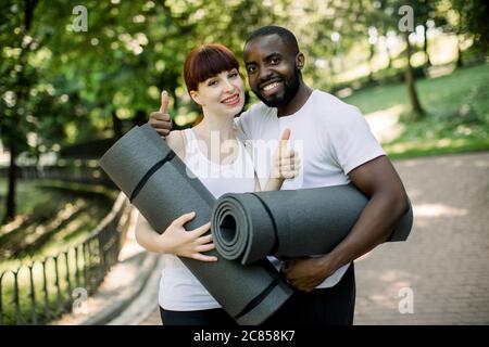 Junge lächelnde multiethnische Paar, afrikanischen Jungen und kaukasischen Mädchen, trägt Sportkleidung, hält Yogamatten, ist bereit für die Ausübung im Stadtpark Stockfoto