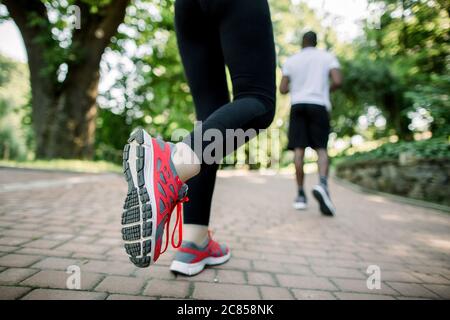 Abgeschnittene Rückansicht eines nicht erkennbaren multiethnischen, athletischen Paares, das im sonnigen Sommerpark läuft und sich aufwärmt. Konzentriere dich auf die Sneaker am weiblichen Bein Stockfoto