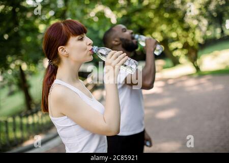 Wasserpause, Sport und Fitness-Konzept. Glückliche junge multiethnische Freunde, kaukasische Frau und afrikanischen Mann, Trinkwasser nach dem Joggen im Park. Seite Stockfoto