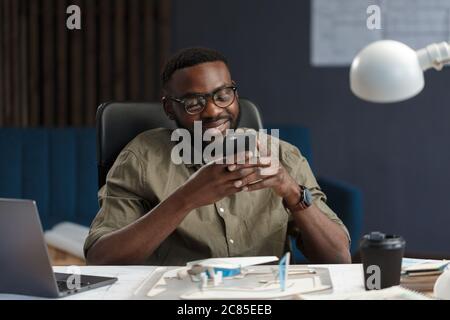 Junger afro-amerikanischer Mann mit Smartphone und Lächeln. Glücklicher Geschäftsmann, der Apps für Mobiltelefone verwendet, Nachrichten getextet, im Internet stöbert, schaut nach Stockfoto