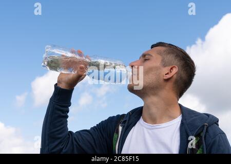 Junger sportlicher Mann trinkt Wasser aus der Flasche gegen blauen Himmel an sonnigen Tagen Stockfoto