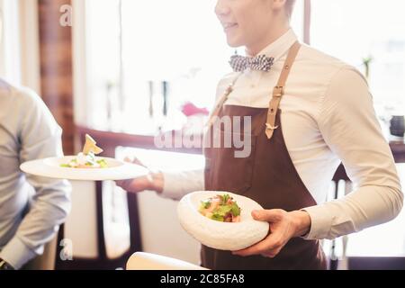 Positiver junger Kellner, der Gäste des Terrassenrestaurants am Tisch serviert. Stockfoto