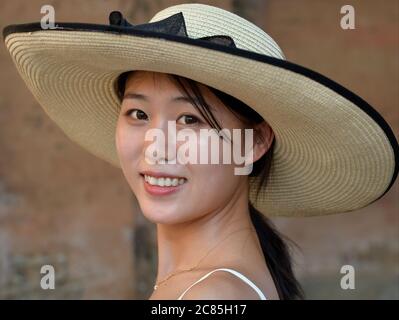 Junge attraktive chinesische Frau im Urlaub trägt einen stilvollen breit-Krempe Boater Strohhut und lächelt für die Kamera. Stockfoto