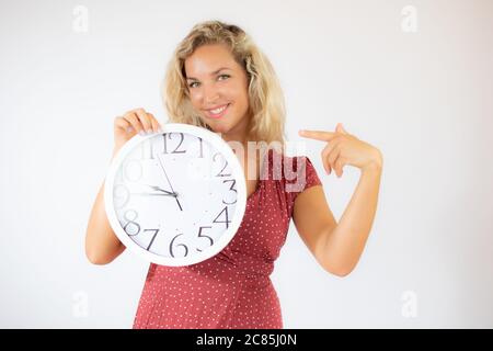 Ziemlich lächelnde blonde Frau in rotem Kleid zeigt eine große Uhr Stockfoto