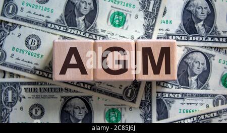 Holzwürfel mit Akronym "AGM" - "Jahreshauptversammlung" auf einem schönen Hintergrund von Dollarscheinen. Geschäftskonzept, Kopierbereich. Stockfoto