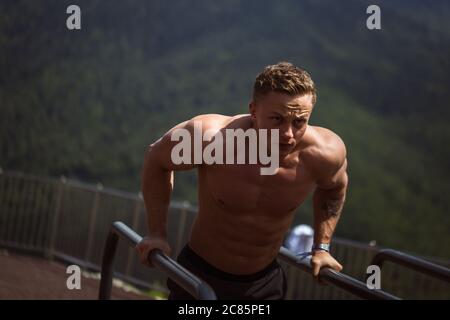 Junge blonde männliche Athlet mit fokussierten und angespannten Gesichtsausdruck die Ausübung im Park tut drücken Sie ups auf parallelen Balken über Berge Hintergrund. Stockfoto